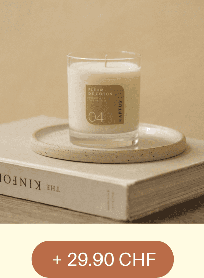 Une bougie blanche en verre étiquetée “FLEUR DE COTON, COLLECTION PRIVÉE, 04, KINTUS” repose sur une assiette. l ensemble est posé sur un livre intitulé 