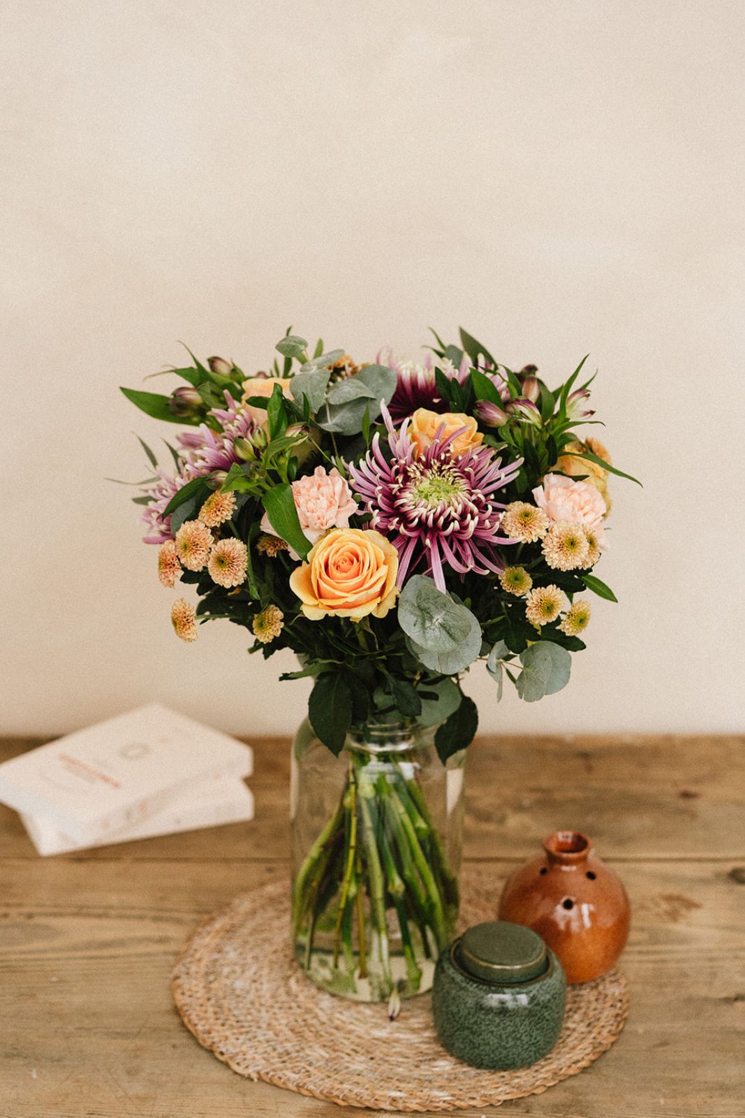 Un bouquet de fleurs variées dans un bocal en verre est posé sur une table en bois, entouré de deux petits pots en céramique et de livres empilés.