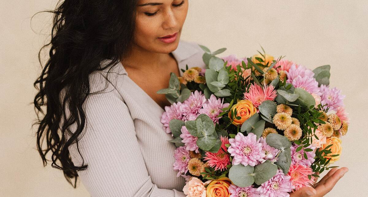 Une femme tient un grand bouquet de fleurs colorées, principalement des roses, chrysanthèmes et dahlias, avec un fond beige uni.