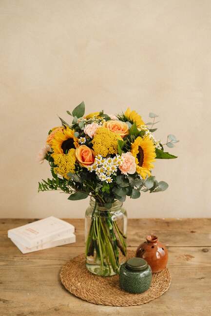 Un bouquet de fleurs, dans un vase en verre, est posé sur une table en bois avec un dessous de plat, entouré de livres et de pots décoratifs en céramique.