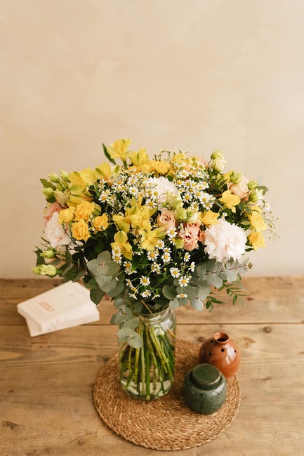 Un vase de fleurs colorées repose sur une table en bois, accompagné d un pot en céramique et d un livre fermé sur un napperon tressé.