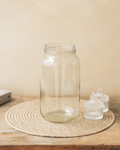 Un pot en verre transparent repose sur un tapis en corde beige, entouré de deux petits pots en verre avec couvercles, le tout posé sur une table en bois.