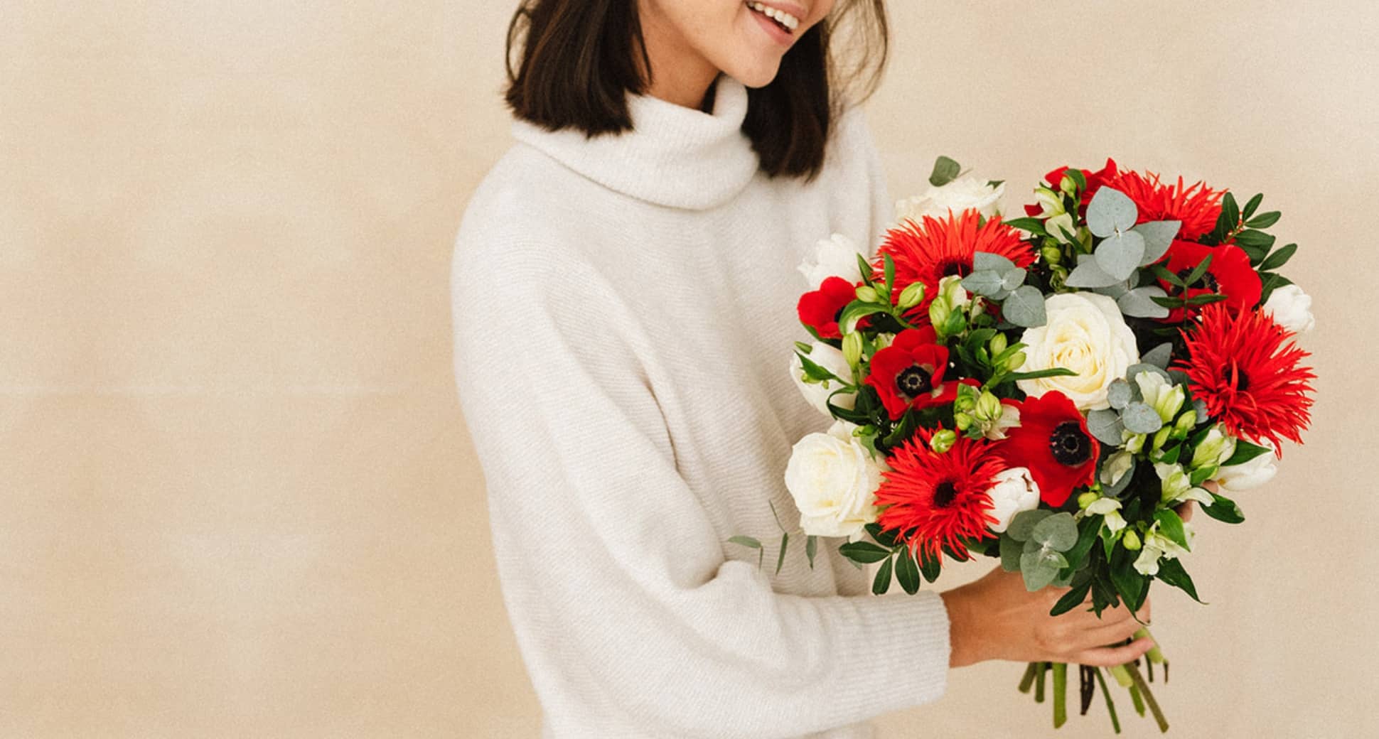 Une personne souriante tient un bouquet de fleurs rouges et blanches dans ses bras, habillée d un pull à col roulé beige, devant un fond beige uni.