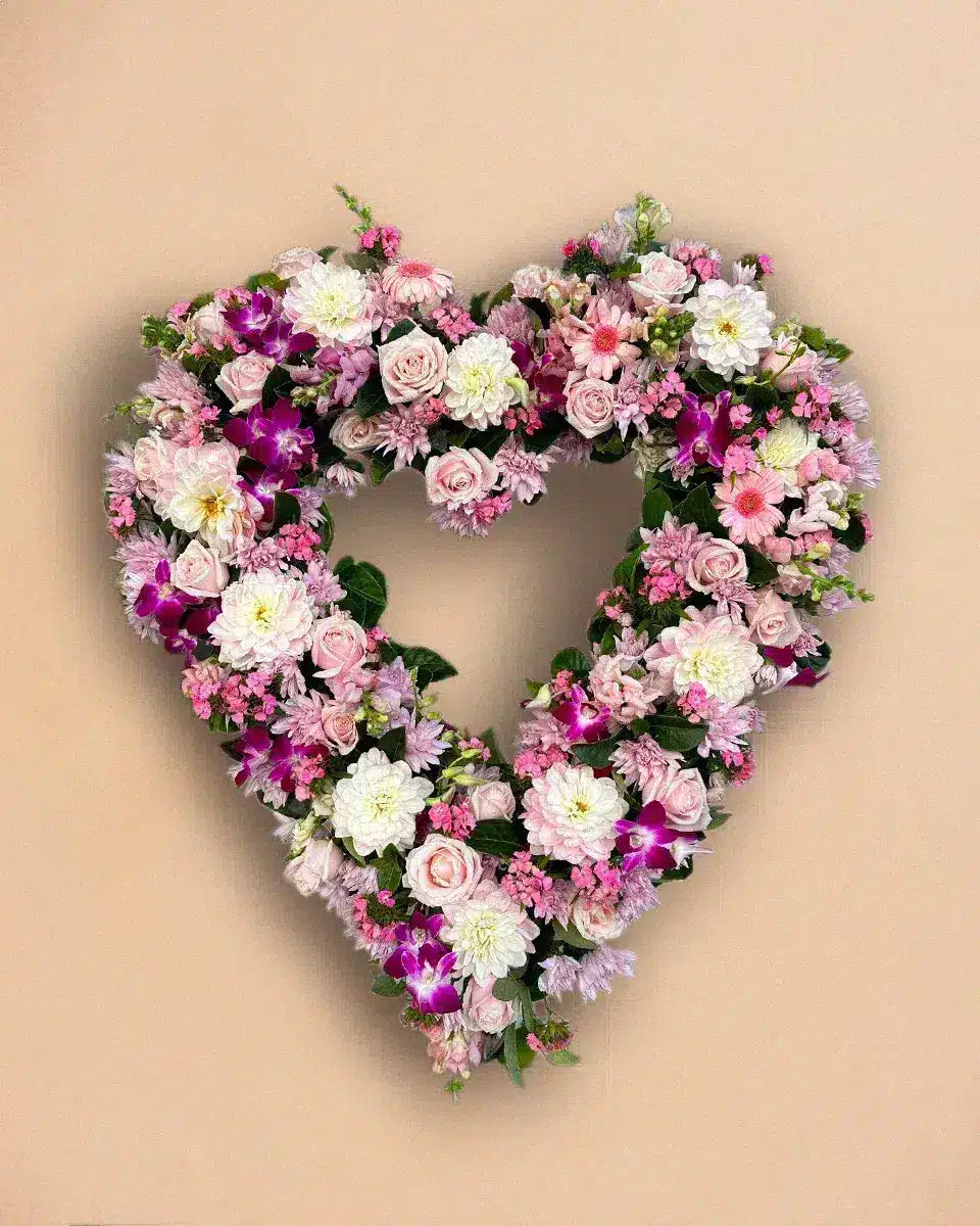 Livraison fleur de deuil : Un bouquet de fleurs colorées disposées en forme de cœur, composé de roses, chrysanthèmes et autres fleurs, suspendu contre un mur beige.