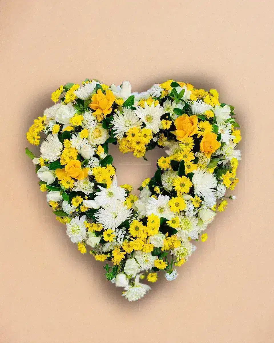 Livraison fleur de deuil : Un cœur de fleurs composé de marguerites blanches, roses jaunes et verdure, suspendu sur un fond beige.