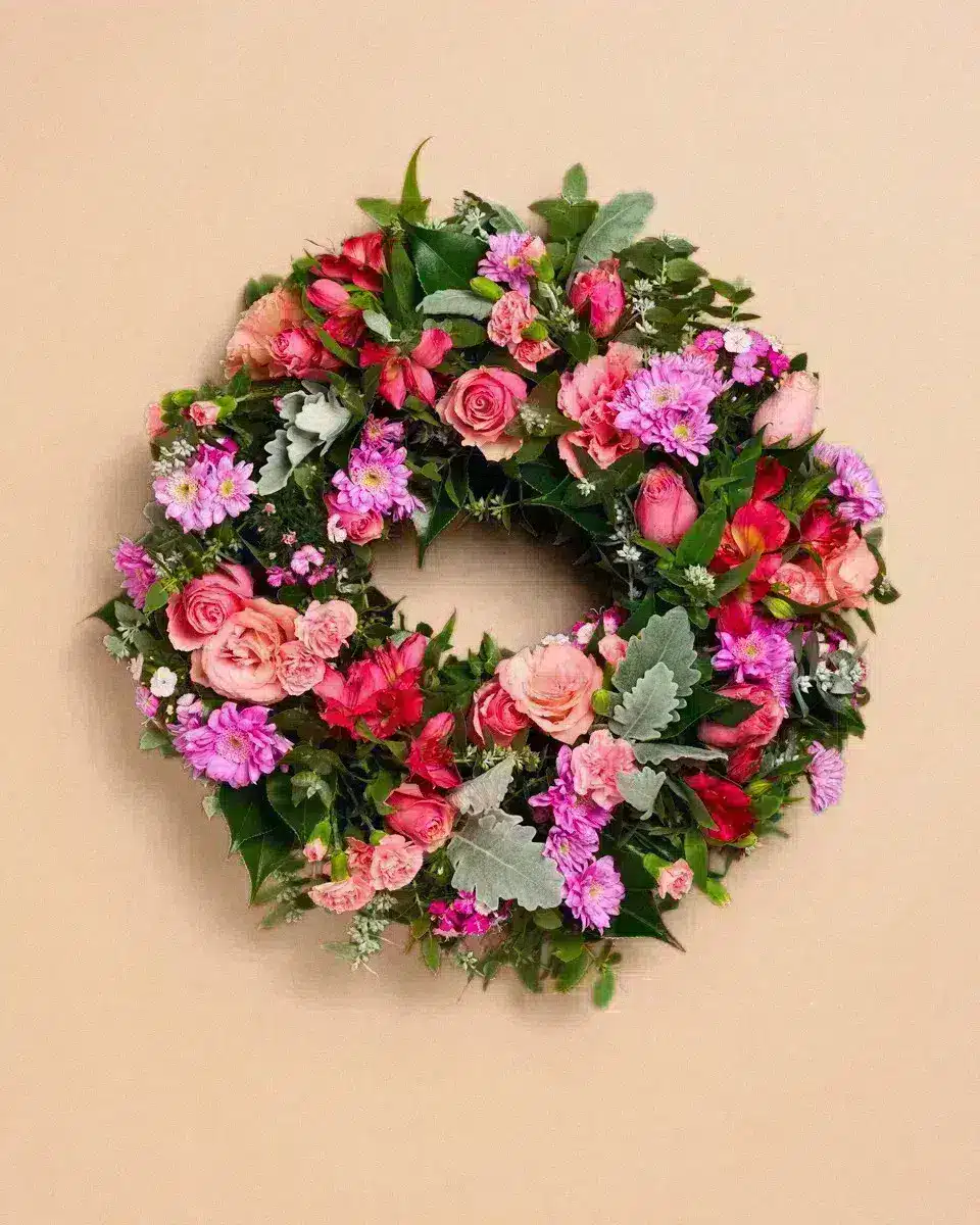 Livraison fleur deuil : Couronne de fleurs colorée ornée de roses, marguerites et feuillage vert, accrochée à un mur beige.