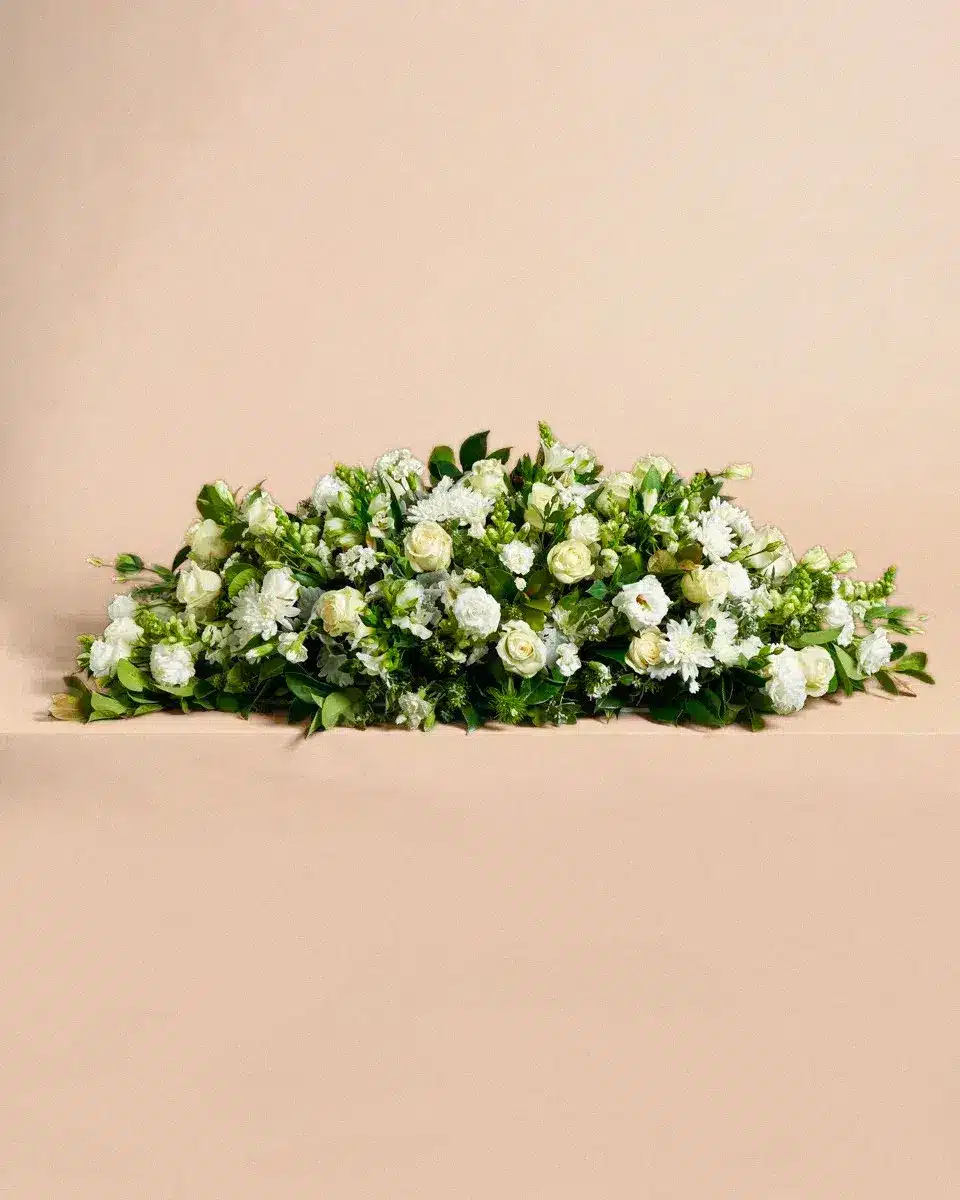 Livraison gerbe de fleur : Composition florale de roses blanches et de feuillage vert, disposée de manière allongée, sur un fond beige neutre.