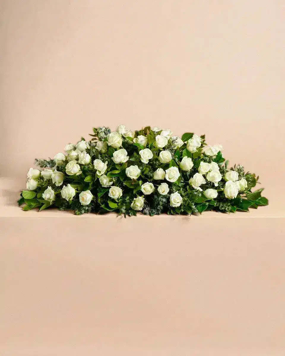 Livraison gerbe de fleur : Un bouquet de roses blanches repose sur une surface beige, entouré de feuillage vert. Il est soigneusement arrangé pour une présentation élégante.