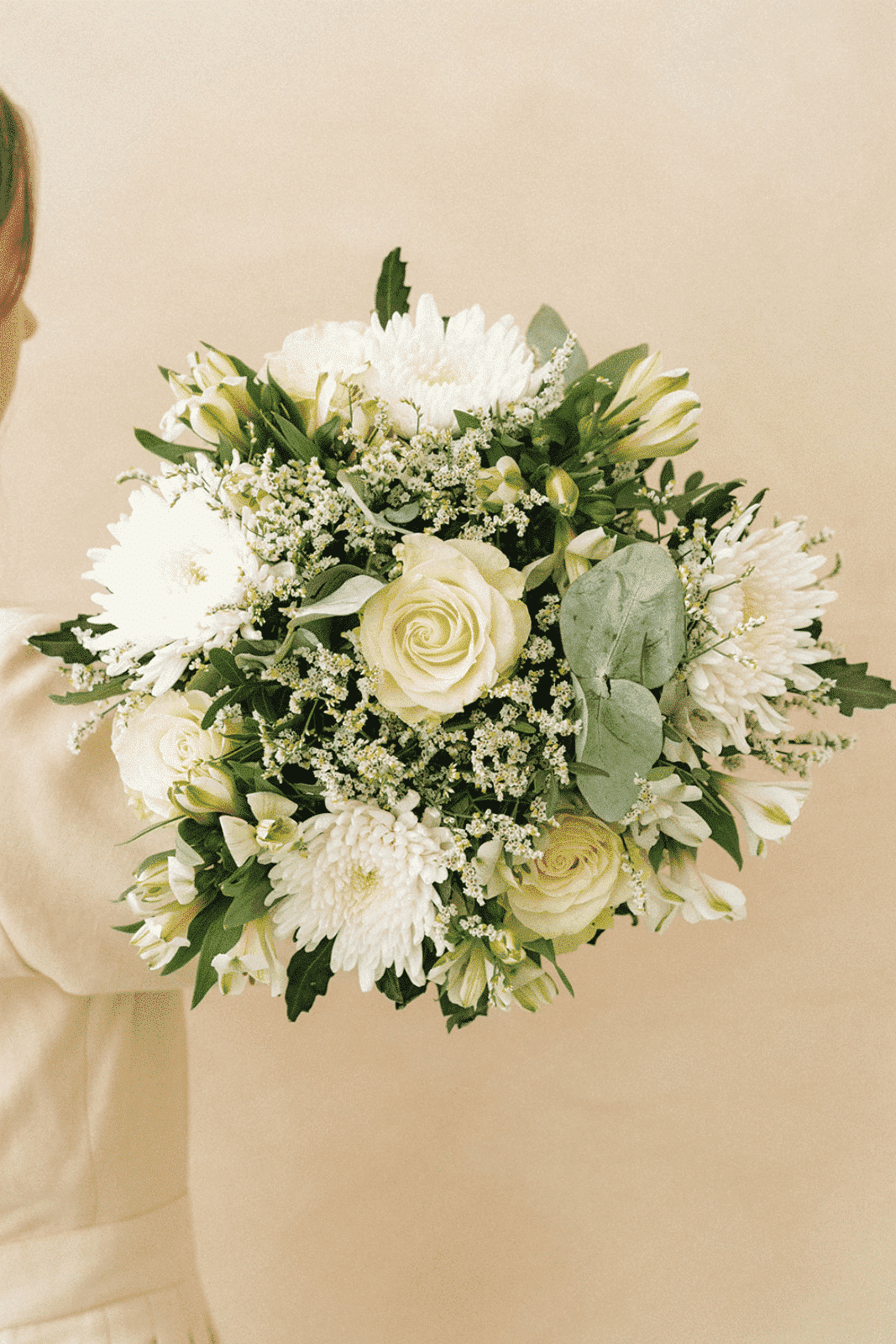 Un bouquet de fleurs blanches et crème, tenu par une personne vêtue de beige, repose sur un fond beige.