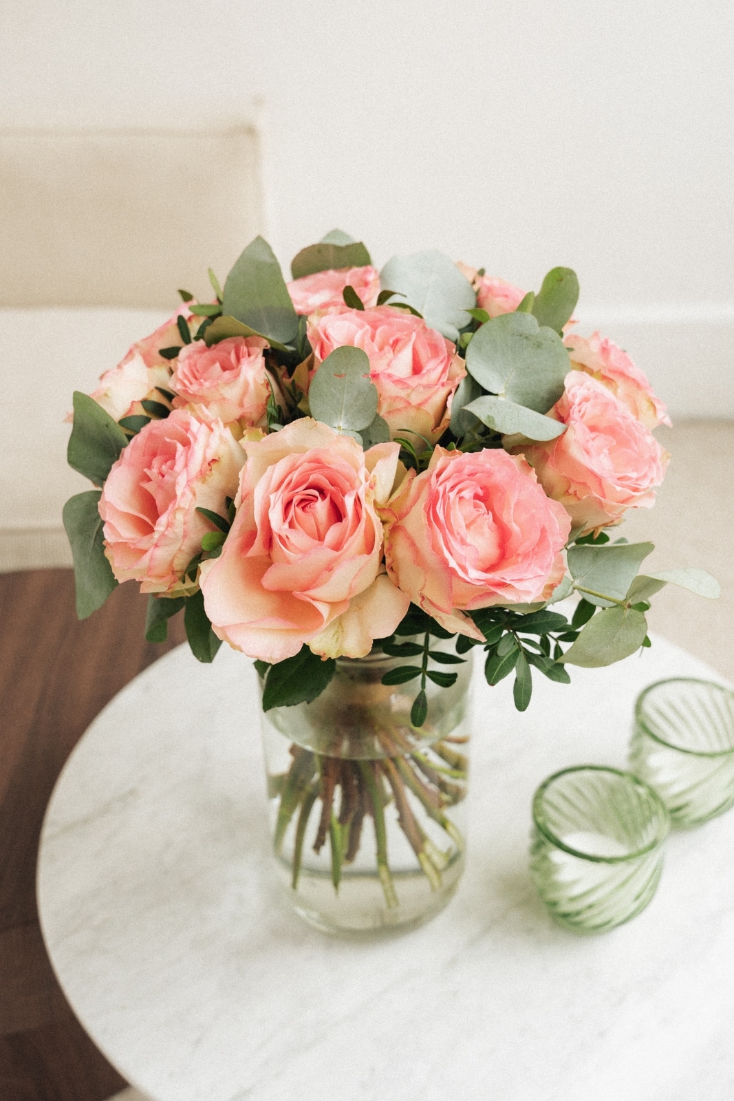 Bouquet de roses roses dans un vase en verre, posé sur une table en marbre, avec des verres verts à proximité, dans une pièce au mobilier clair.