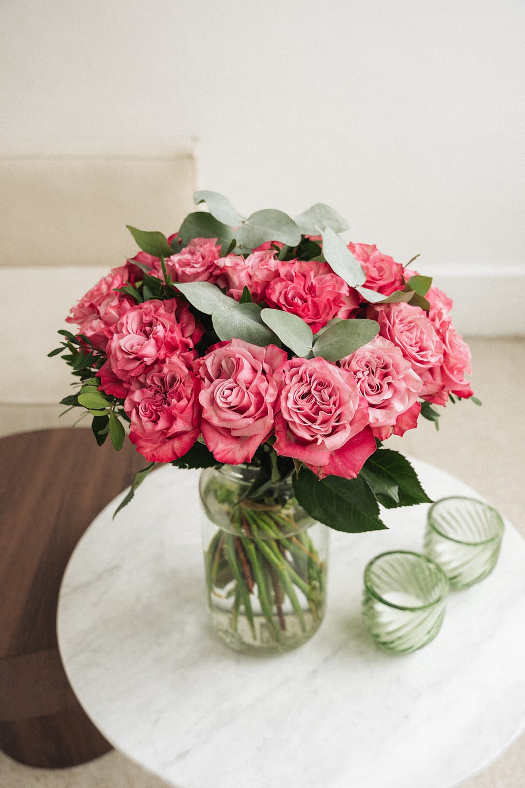 Ein Strauss rosafarbener Rosen steht in einer durchsichtigen Glasvase auf einem Marmortisch neben zwei strukturierten grünen Gläsern in einem hellen, minimalistischen Raum.