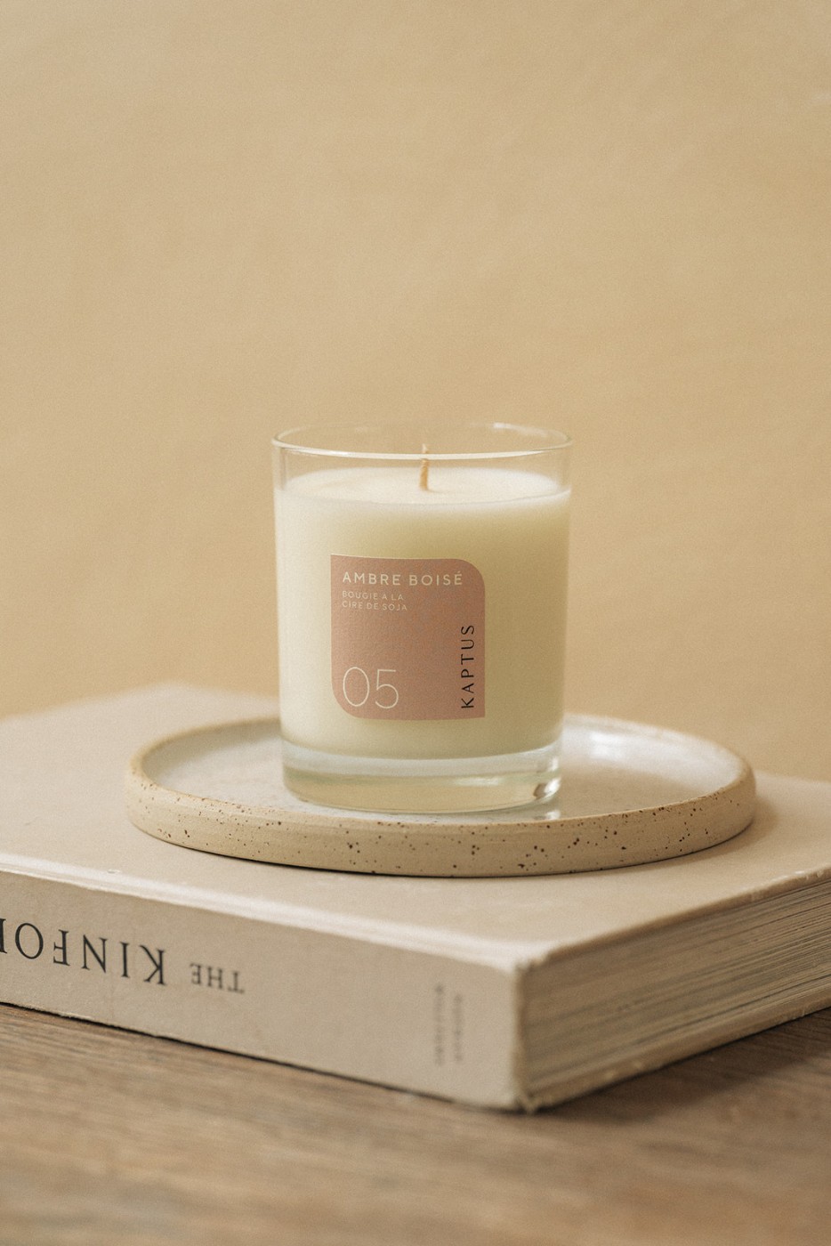 Bougie parfumée dans un verre transparent, posée sur une soucoupe beige, elle-même placée sur le livre