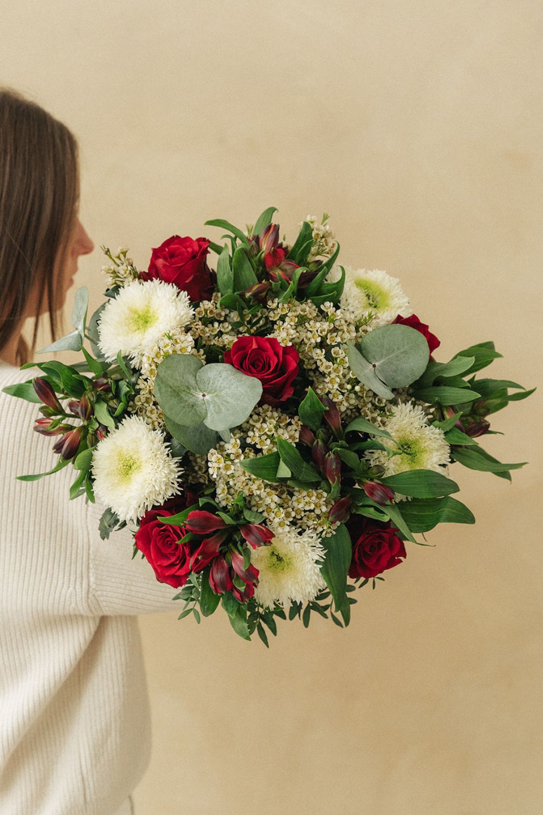Un bouquet de roses rouges, de chrysanthèmes blancs et de verdure est tenu par une personne vêtue de blanc, de profil, sur un fond beige uni.