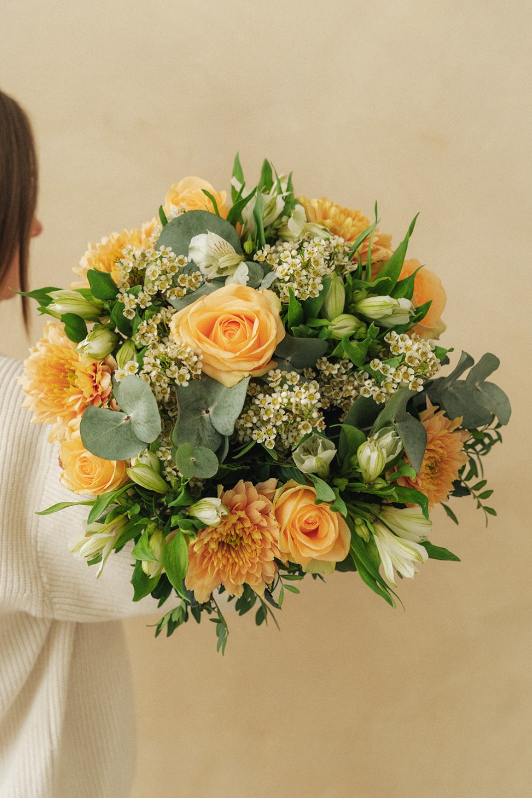 Un bouquet de fleurs jaunes et blanches, comprenant des roses et des chrysanthèmes, tenu par une personne en pull crème contre un fond beige.